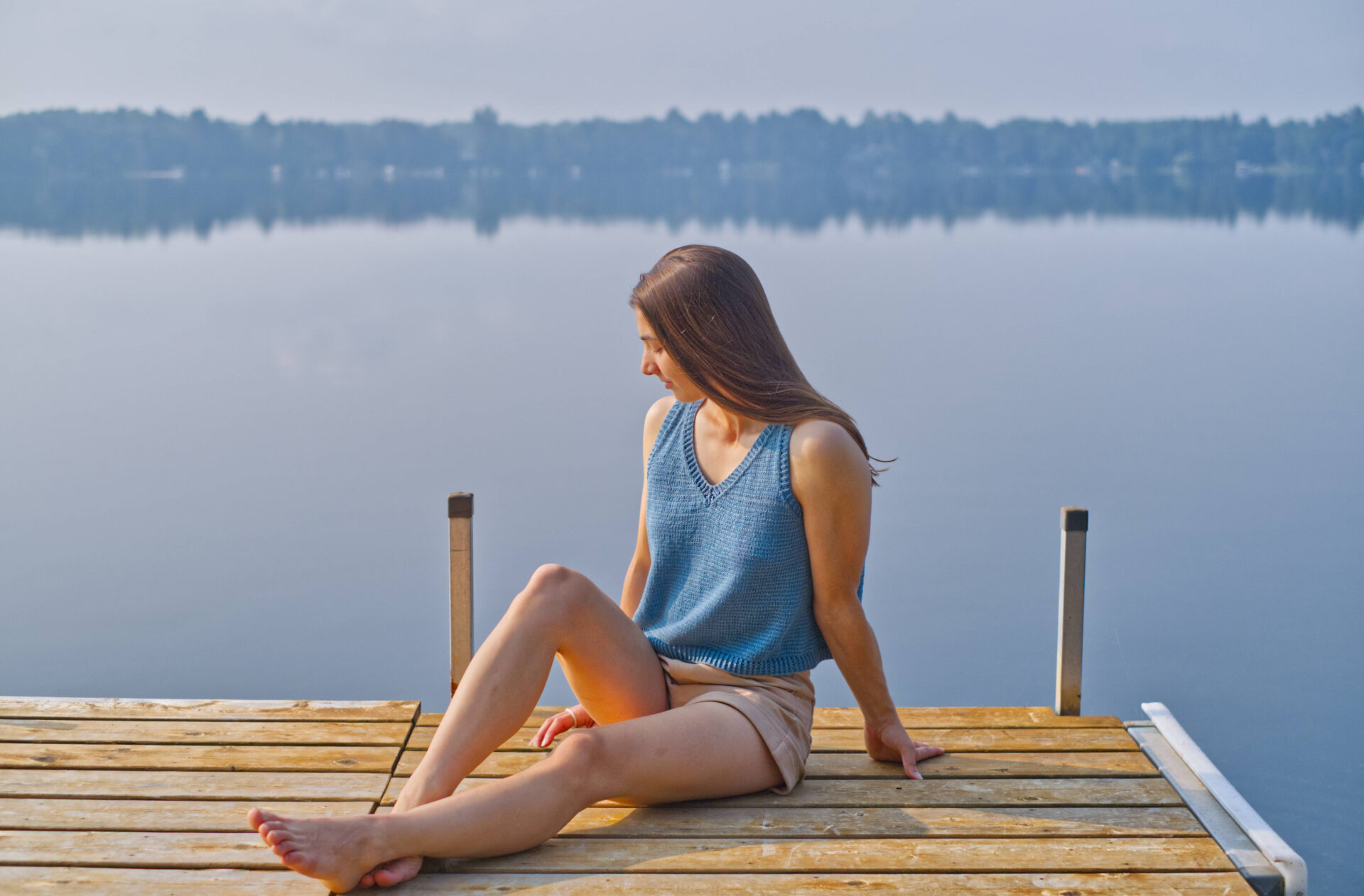 shena is lounging on the dock in front of a calm blue lake, wearing a blue v neck knit top.